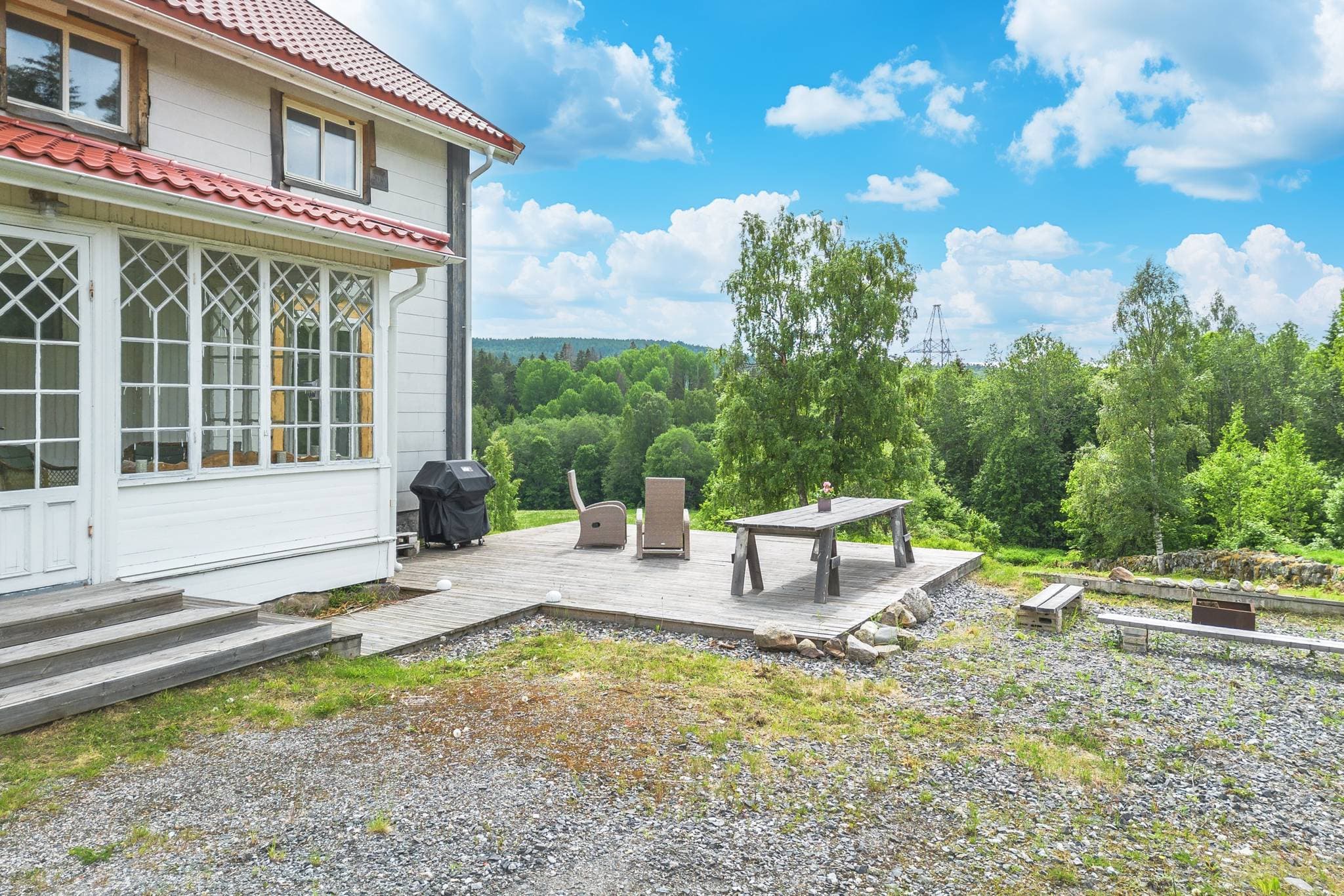 Övre bostadshusets altan och eldplats med utsikt över gräsmatta, åker, stenmur och skog. Riktning syd