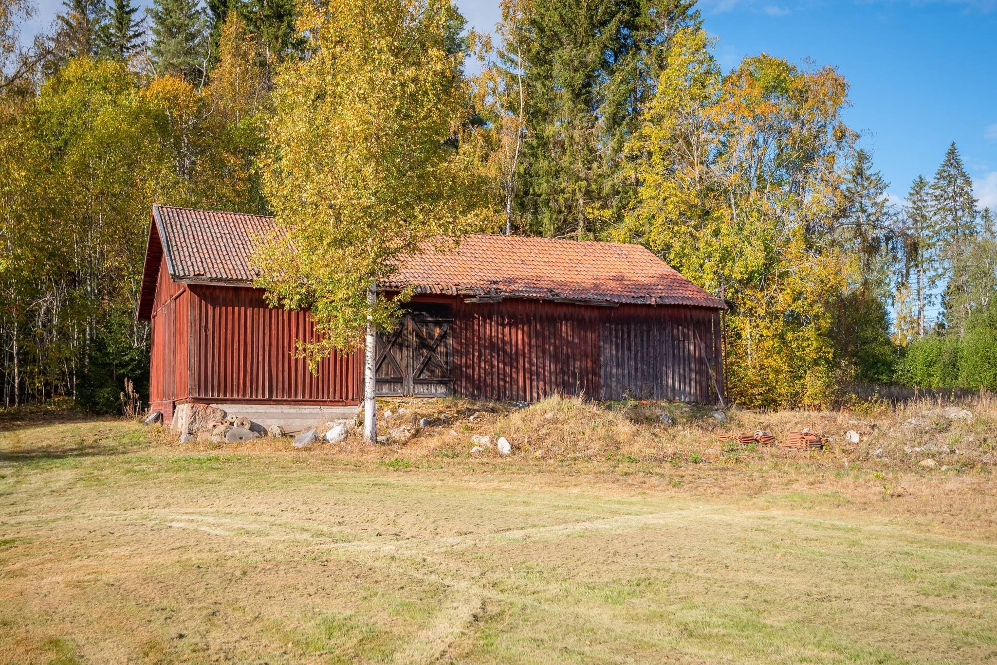 Förrådsbyggnad Denna välhållna ekonomibyggnad, uppförd under början av 1900-talet, erbjuder praktiska och flexibla förvaringsmöjligheter. Byggnaden moderniserades under 1980-talet och har idag ett gjutet betonggolv som ger en slitstark och lättskött yta. Byggnaden är oisolerad, välventilerad och håller tätt, vilket skapar goda förutsättningar för säker förvaring året om. Den stora skjutdörren på gaveln tillsammans med dörren på långsidan gör byggnaden lättillgänglig och smidig att använda för såväl maskiner och redskap som material och annan utrustning. Med sitt strategiska läge i gårdsmiljön och sin funktionella utformning är detta en byggnad som enkelt kan anpassas efter olika behov, oavsett om den används som förråd, verkstad eller för annan verksamhet.